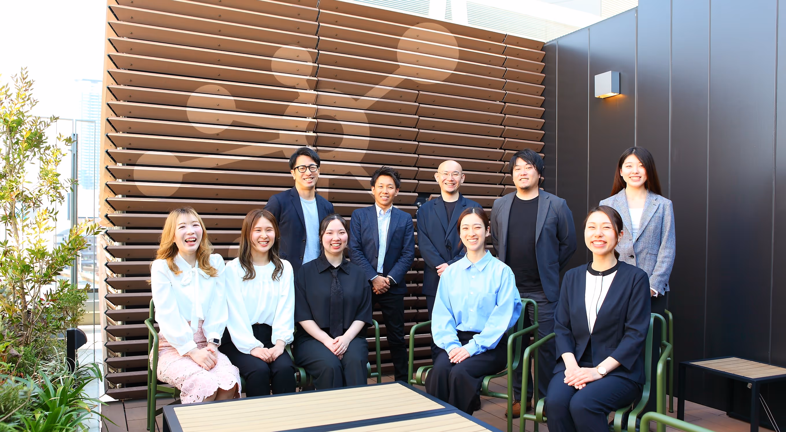 Group of nine people, five women seated and four men standing behind them, smiling in an outdoor patio setting.